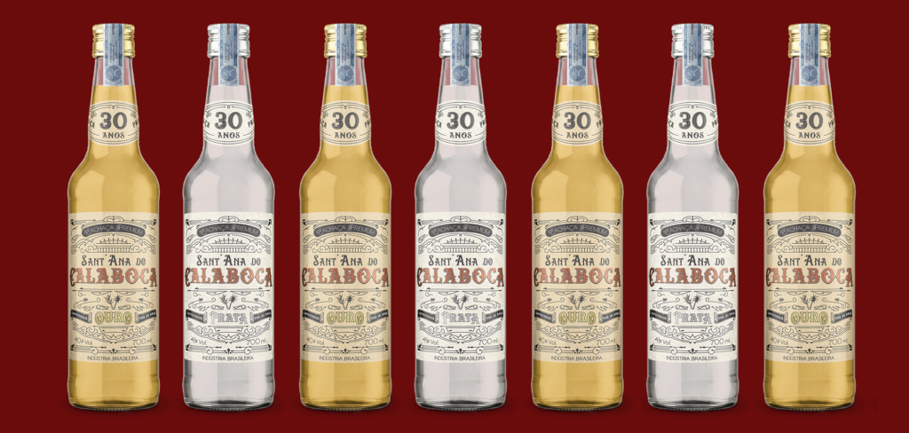 Cinco garrafas da Cachaça Sant'Ana do Calaboca enfileiradas sobre fundo bordô, alternando entre as versões Ouro e Prata. Os rótulos possuem inspiração colonial, tipografia clássica e detalhes que remetem ao estilo barroco.